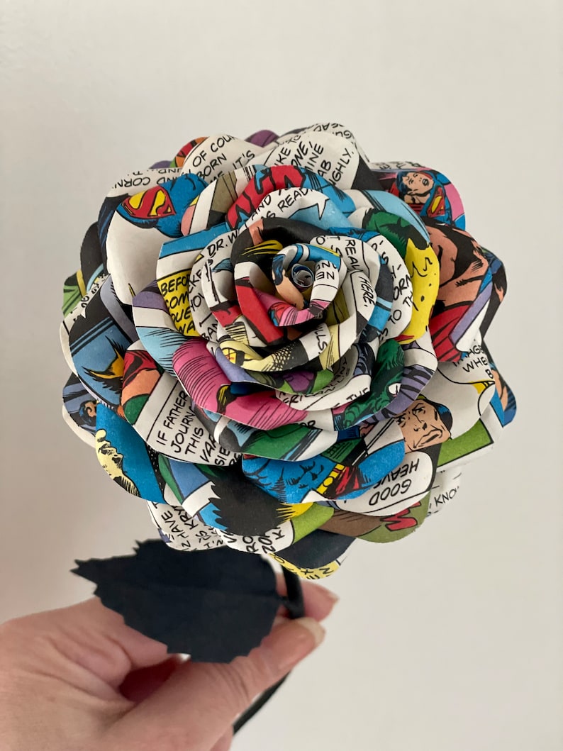 Puede incluir: Una rosa de papel hecha de p&aacute;ginas de c&oacute;mics con Superman, Batman y otros superh&eacute;roes. La rosa es una mezcla vibrante de rojo, azul, amarillo y verde. El tallo es negro.