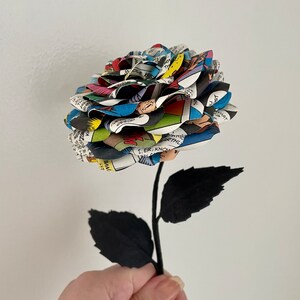 Puede incluir: Un solo tallo negro con dos hojas sostiene una rosa hecha de p&aacute;ginas de c&oacute;mics. La rosa es una mezcla vibrante de colores, incluyendo rojo, azul, amarillo y verde.