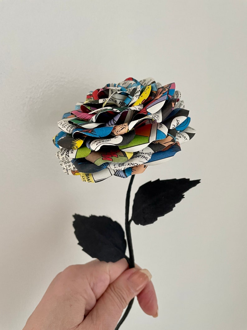 Puede incluir: Un solo tallo negro con dos hojas sostiene una rosa hecha de p&aacute;ginas de c&oacute;mics. La rosa es una mezcla vibrante de colores, incluyendo rojo, azul, amarillo y verde.