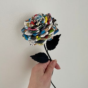 Puede incluir: Un solo tallo negro con hojas negras sostiene una rosa hecha de p&aacute;ginas de c&oacute;mics. La rosa es multicolor y presenta una variedad de personajes de c&oacute;mics.