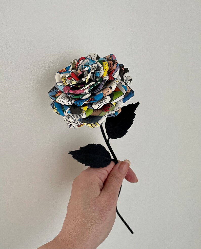 Puede incluir: Un solo tallo negro con hojas negras sostiene una rosa hecha de p&aacute;ginas de c&oacute;mics. La rosa es multicolor y presenta una variedad de personajes de c&oacute;mics.
