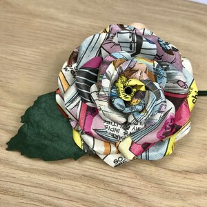 May include: A colorful paper rose made from comic book pages. The rose is pink, yellow, blue, and white. It has a green paper leaf.