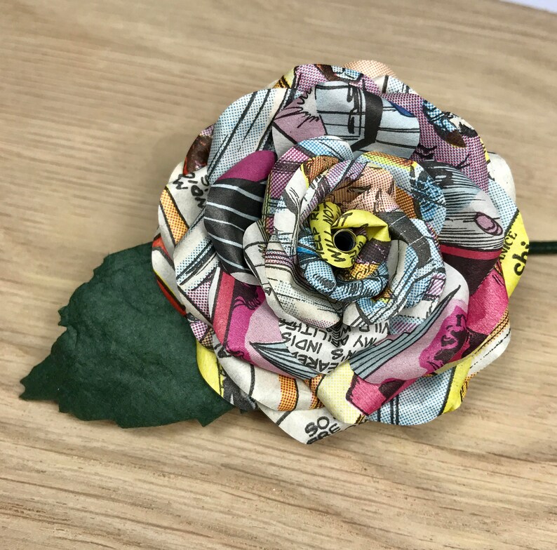 May include: A colorful paper rose made from comic book pages. The rose is pink, yellow, blue, and white. It has a green paper leaf.
