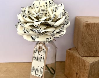 Puede incluir: Una rosa blanca de papel hecha de partituras musicales se muestra en una botella de vidrio transparente con una etiqueta de papel marrón. La etiqueta tiene notas musicales impresas. La rosa está sobre una superficie de madera.
