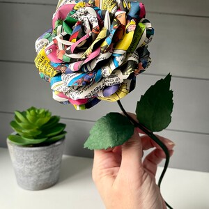 Puede incluir: Una rosa de papel colorida hecha de p&aacute;ginas de c&oacute;mics. La rosa es una mezcla vibrante de rojo, amarillo, azul, verde y negro. El tallo es verde y tiene dos hojas.
