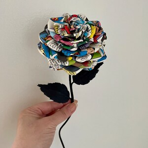 Puede incluir: Una sola rosa de c&oacute;mic con tallo y hojas negras. La rosa est&aacute; hecha de p&aacute;ginas de un c&oacute;mic con ilustraciones y texto coloridos.