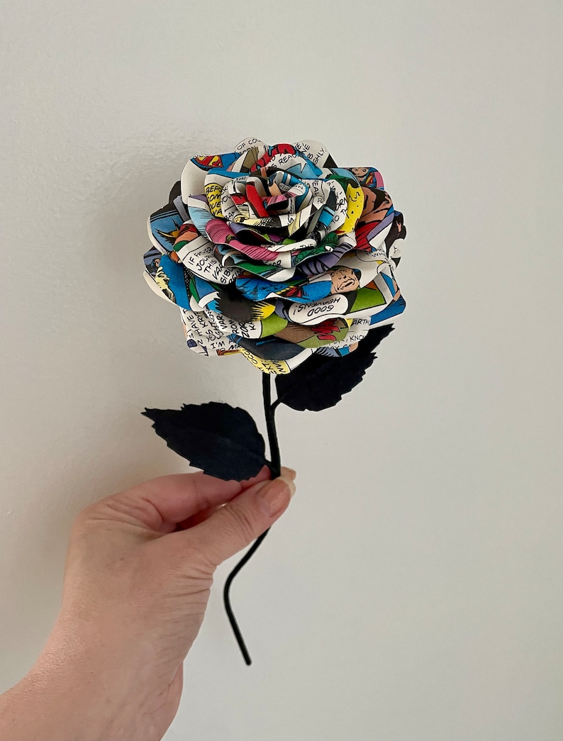 Puede incluir: Una sola rosa de c&oacute;mic con tallo y hojas negras. La rosa est&aacute; hecha de p&aacute;ginas de un c&oacute;mic con ilustraciones y texto coloridos.