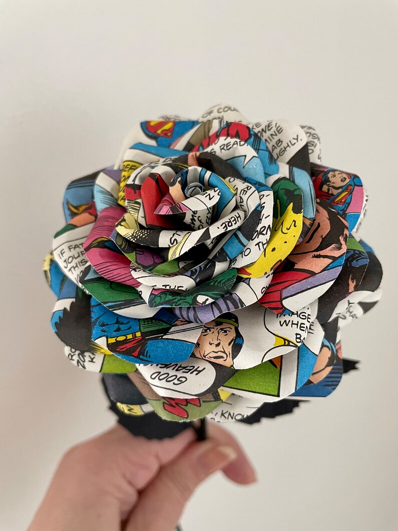 Puede incluir: Una rosa de tallo negro hecha de p&aacute;ginas de c&oacute;mics con Superman. La rosa est&aacute; hecha de m&uacute;ltiples capas de p&aacute;ginas de c&oacute;mics, con el texto "HEAVEN GOOD" visible en una de las capas.
