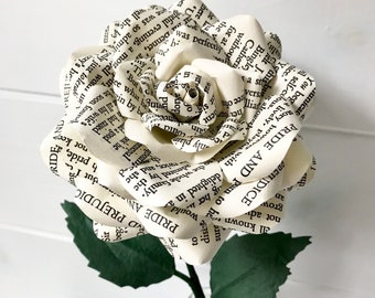 May include: A white paper rose made from the pages of the book "Pride and Prejudice". The rose is in a glass vase with green leaves.