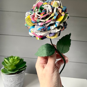 Puede incluir: Una rosa de papel colorida hecha de p&aacute;ginas de c&oacute;mics. La rosa es una mezcla vibrante de rojo, amarillo, azul, verde y negro. El tallo es verde y tiene dos hojas.