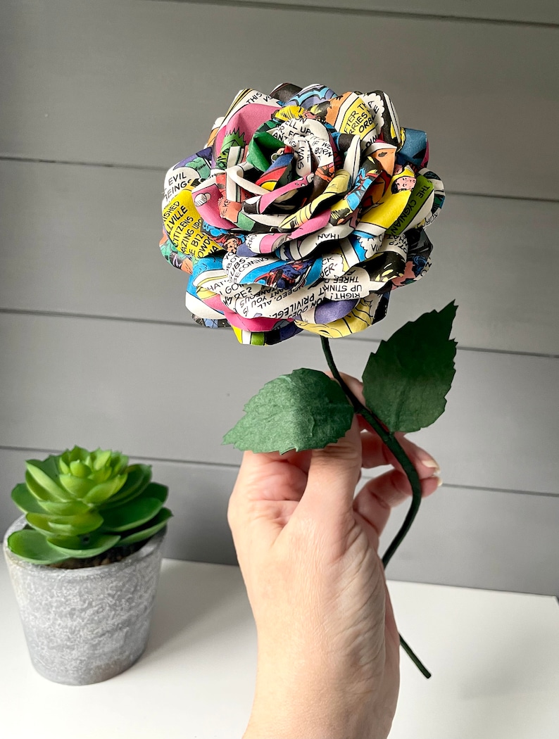 Puede incluir: Una rosa de papel colorida hecha de p&aacute;ginas de c&oacute;mics. La rosa es una mezcla vibrante de rojo, amarillo, azul, verde y negro. El tallo es verde y tiene dos hojas.
