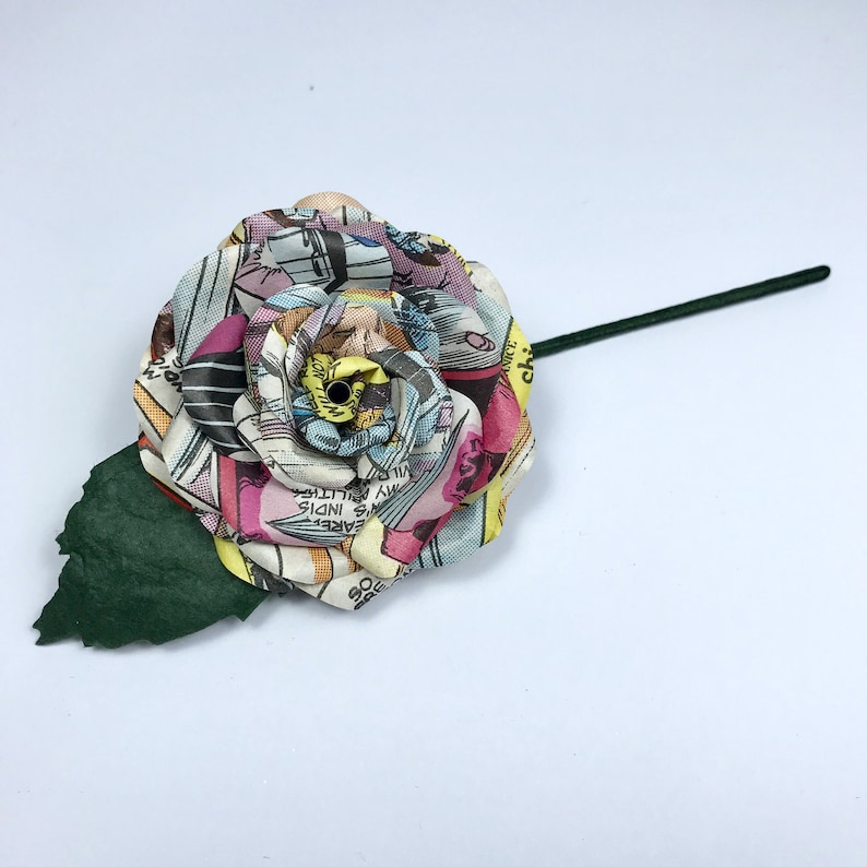 May include: A single, handmade paper rose made from recycled comic book pages. The rose is a light pink, yellow, and blue color with a green leaf. The rose is on a white background.