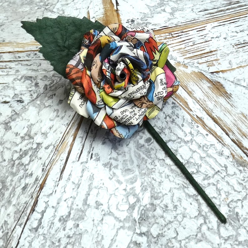 May include: A colorful comic book rose with a green stem and leaf. The rose is made of fabric and has a vintage comic book pattern. The text on the comic book fabric reads "KEEPING YOU", "CUT", "DIDN'T", "AN STRENG", "AN TONIG", "NA USE 'E", "REEPING", "YOU", "S."