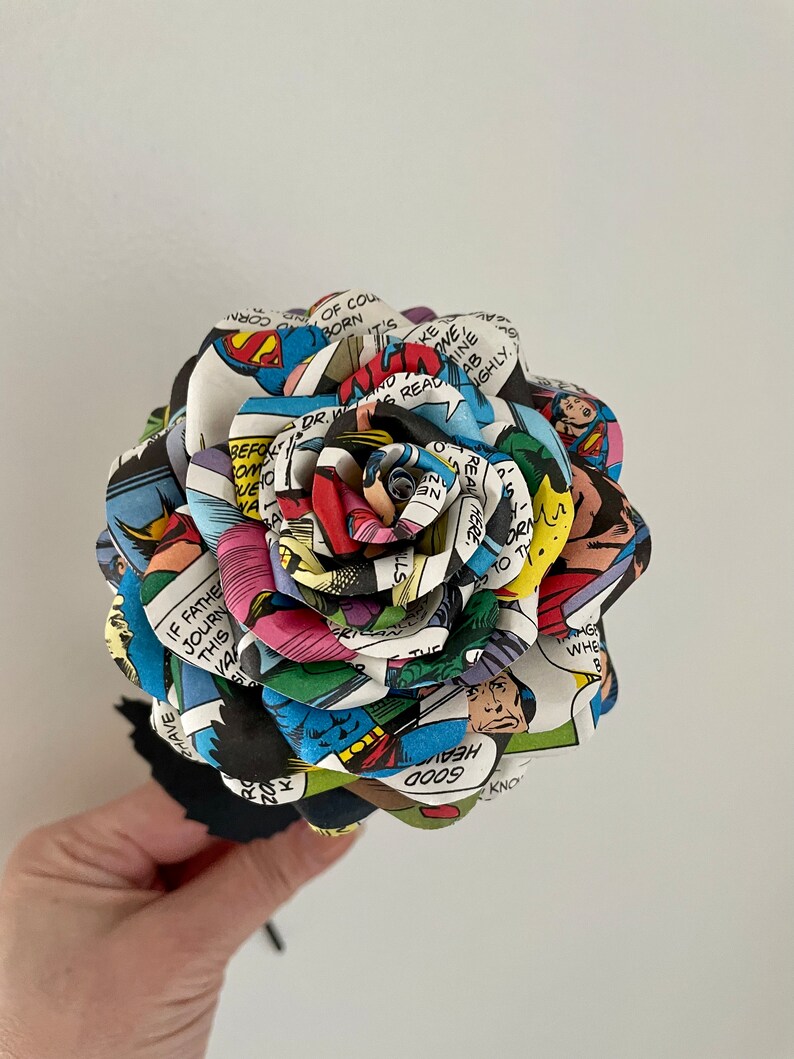 Puede incluir: Una rosa de papel colorida hecha de p&aacute;ginas de c&oacute;mics. La rosa est&aacute; hecha de diferentes paneles de colores de c&oacute;mics, incluyendo Superman, Batman y otros superh&eacute;roes. La rosa se sostiene en una mano con un tallo negro.
