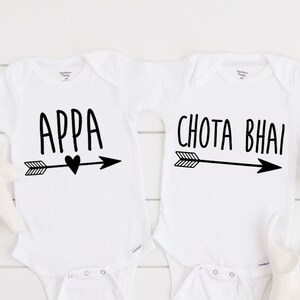 May include: Two white baby onesies with black text and arrows. The onesie on the left says "Appa" with a heart above the arrow. The onesie on the right says "Chota Bhai".