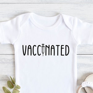 May include: White baby onesie with black text that reads "VACCINATED" with a syringe graphic above the word "VACCINATED".