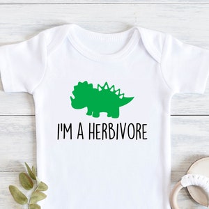 May include: White baby onesie with a green dinosaur graphic and the text "I'M A HERBIVORE". The onesie is short-sleeved and has a simple, clean design. A wooden teething ring and a sprig of greenery are next to the onesie.