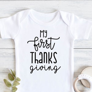 Puede incluir: Un body blanco para bebé con texto negro que dice "My first thanks giving".