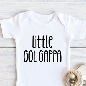 May include: White baby onesie with the text "little GOL GAPPA" in black. A wooden and cream-colored baby rattle and a small wooden disc are next to the onesie. The onesie is on a white wooden surface.