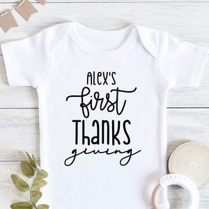 Könnte beinhalten: Weißer Baby-Body mit dem schwarzen Schriftzug "Alex's first Thanksgiving". Das Bild zeigt einen Beißring aus Holz, einen Zweig mit Grün und kleine Wimpel-Fahnen in Beige als dekorative Elemente.