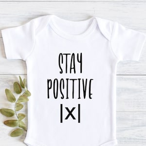 Op de afbeelding: Witte baby romper met zwarte tekst die "STAY POSITIVE [X]" luidt.