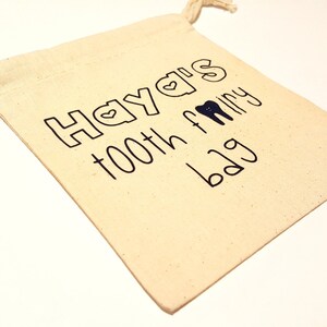 Könnte beinhalten: Ein kleiner, weißer Beutel mit Kordelzug, auf dem der Text "Hayla's tooth fairy bag" in Schwarz aufgedruckt ist. Der Text enthält ein kleines Herz und eine Zahnzeichnung.