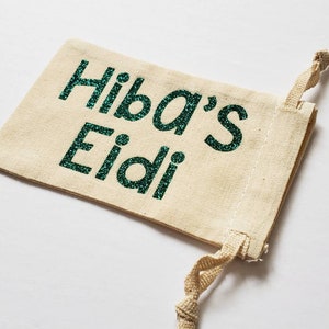 Può includere: Un piccolo sacchetto beige con coulisse con le parole "Hiba's Eidi" scritte in glitter verde.