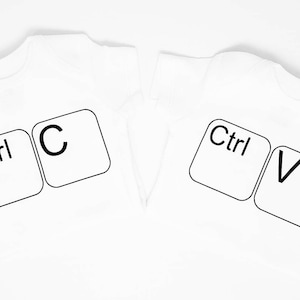 以下が含まれることがあります： 黒いコンピューターキーボードのキーが印刷された白いTシャツが2枚。最初のシャツには「Ctrl」と「C」のキーが、2番目のシャツには「Ctrl」と「V」のキーが印刷されています。