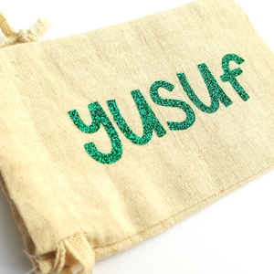 May include: A small, beige drawstring bag with a green glitter design that says "YUSUF".