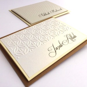 May include: Two white greeting cards with brown envelopes. The top card has a white background with small white dots and the text "Nikkah Mubarak". The bottom card has a white background with a white embossed pattern and the text "Jazak Allah".