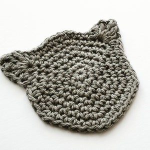 May include: A gray crocheted cat-shaped coaster. The coaster is made of a soft, textured yarn and has a simple, minimalist design.