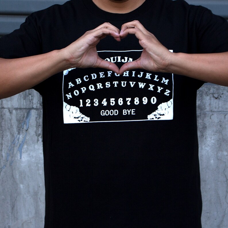 Puede incluir: Camiseta negra con un gr&aacute;fico blanco de una tabla Ouija con las letras A-Z, los n&uacute;meros 1-9 y 0, y las palabras "Good Bye".