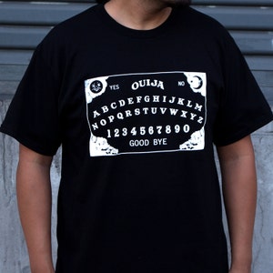 Puede incluir: Camiseta negra con un gr&aacute;fico blanco de una tabla Ouija. La tabla incluye las letras A-Z, los n&uacute;meros 0-9 y las palabras "YES", "NO" y "GOOD BYE".