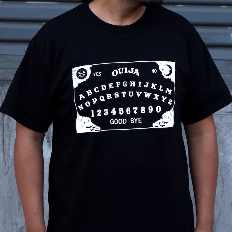Puede incluir: Camiseta negra con un gr&aacute;fico blanco de una tabla Ouija. La tabla incluye las letras A-Z, los n&uacute;meros 0-9 y las palabras "YES", "NO" y "GOOD BYE".