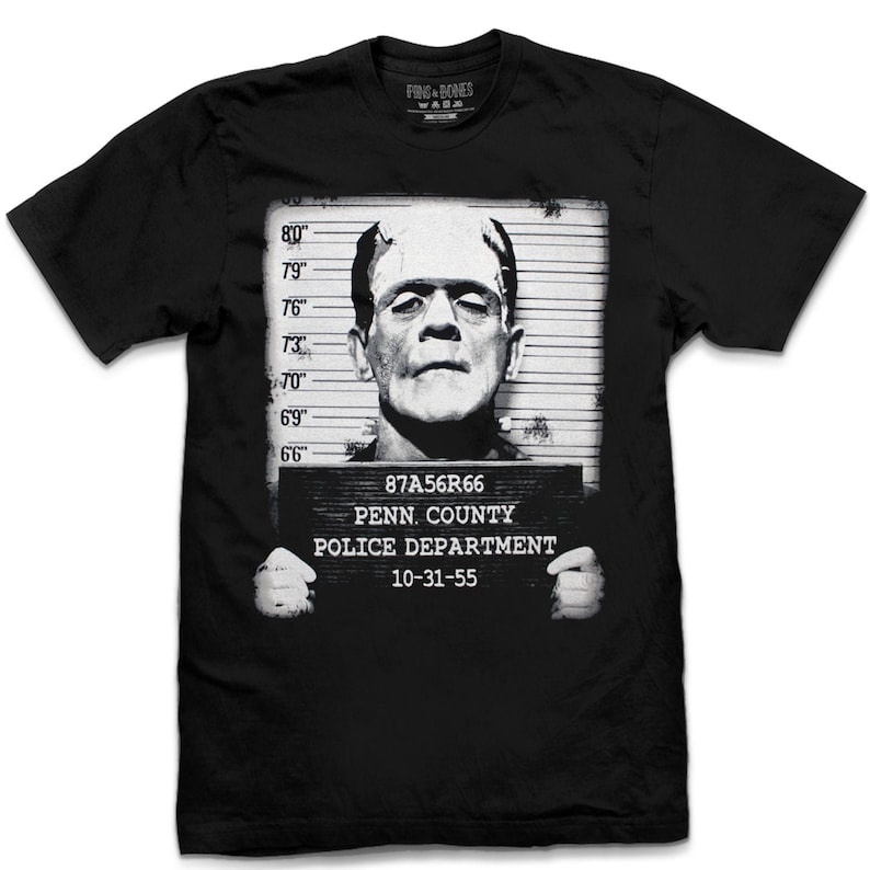 May include: Black t-shirt with a white graphic of Frankenstein's monster in a mugshot. The graphic includes the text "87A56R66", "PENN. COUNTY", "POLICE DEPARTMENT", and "10-31-55".