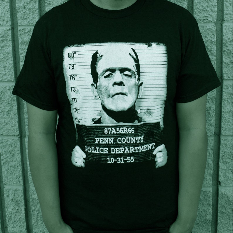 May include: Black t-shirt with a graphic print of Frankenstein in a mugshot. The text on the mugshot reads "87A56R66 PENN. COUNTY POLICE DEPARTMENT 10-31-55".