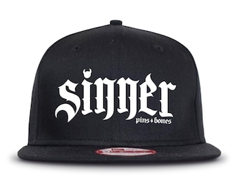 Puede incluir: Gorra de béisbol negra y blanca con la palabra "Sinners" en fuente gótica y el texto "pins + bones" debajo.