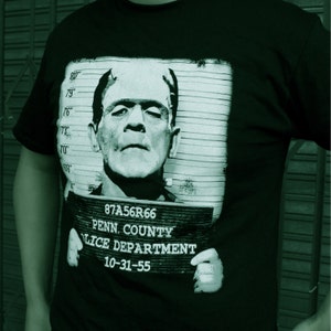 May include: Black t-shirt with a graphic print of a black and white mugshot of Frankenstein. The mugshot includes the text "87A56R66", "PENN. COUNTY", "LICE DEPARTMENT", and "10-31-55".