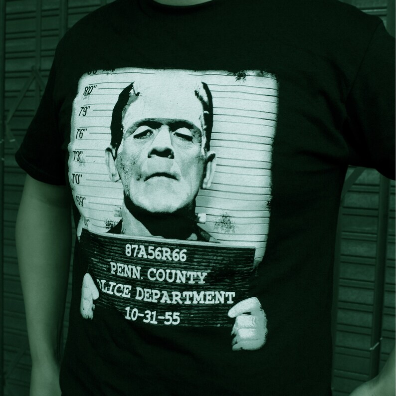 May include: Black t-shirt with a graphic print of a black and white mugshot of Frankenstein. The mugshot includes the text "87A56R66", "PENN. COUNTY", "LICE DEPARTMENT", and "10-31-55".