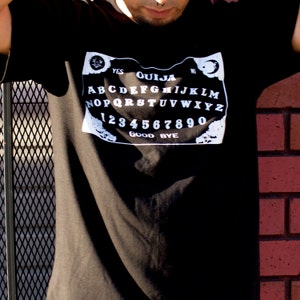 Puede incluir: Una camiseta negra con un gr&aacute;fico blanco de una tabla Ouija. La tabla presenta las letras A-Z, los n&uacute;meros 1-9 y las palabras "YES", "NO", "GOOD BYE" y "OUIJA".
