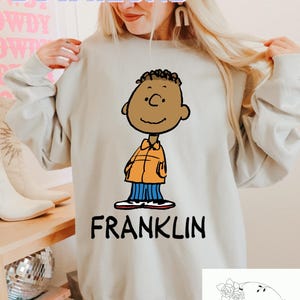 Könnte beinhalten: Ein hellgraues Sweatshirt mit einer Cartoon-Illustration von Franklin aus der Peanuts-Comicserie. Die Illustration ist in Schwarz, Orange und Blau gehalten. Der Text "FRANKLIN" ist in Schwarz unter der Illustration aufgedruckt. Das Sweatshirt wird von einer Person getragen. Das Bild enthält auch ein Logo für "Wildflower Designs" in der unteren rechten Ecke.