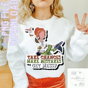 Puede incluir: Sudadera blanca con una ilustración de dibujos animados de una mujer montando un dragón verde. El texto "Take Chances Make Mistakes Get Messy" está impreso en la sudadera.