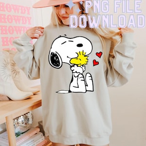 May include: A beige sweatshirt with a graphic of Snoopy kissing Woodstock, and two red hearts. The sweatshirt has long sleeves and a crew neck. Text in the image reads "PNG FILE DOWNLOAD".