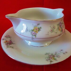 May include: A white porcelain gravy boat and saucer set. The set features a floral pattern with yellow, pink, and purple roses. Gold trim accents the edges of the boat and saucer. A red line encircles the boat.