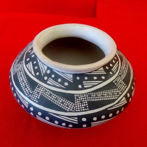 May include: A handcrafted ceramic pot with a rounded body and a wide, open mouth. The pot features a black and white geometric pattern, with a white rim and a red background. The design includes lines, dots, and square patterns.