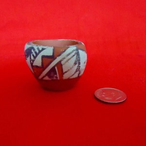 May include: Small, hand-painted ceramic bowl with a geometric design in white, black, and orange. The bowl is round with a slightly flared rim and sits on a red background. A coin is next to the bowl.