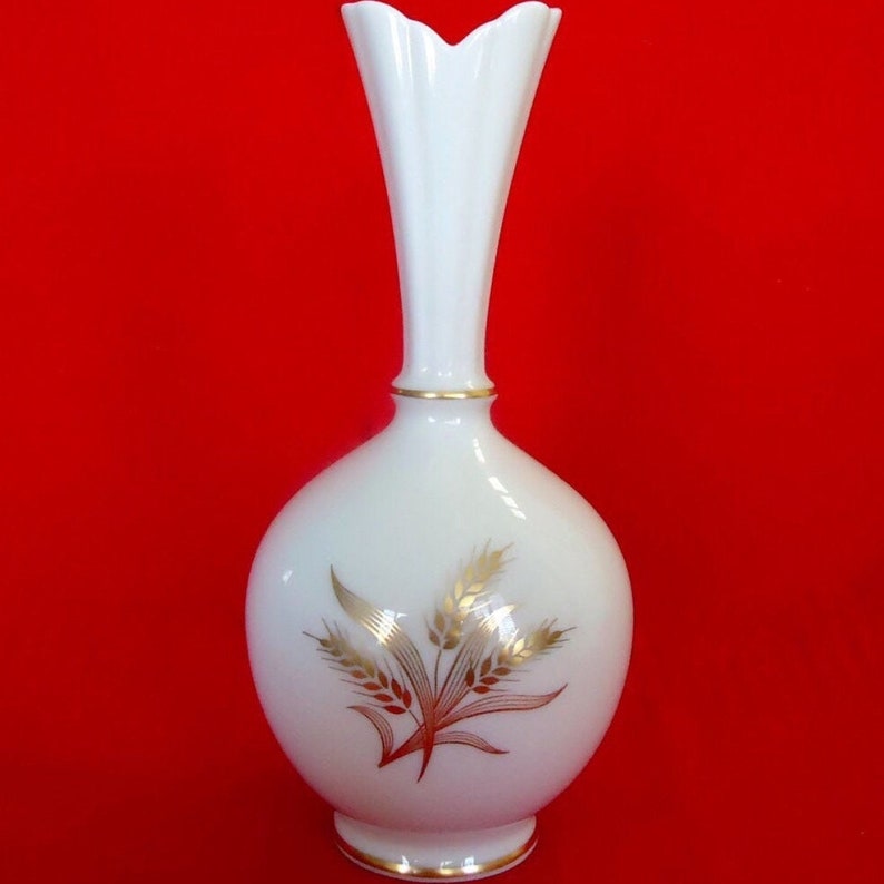 May include: A white ceramic vase with a bulbous base and a slender, fluted neck. The vase features a gold band at the neck and base, and a wheat design in gold and brown. The vase is approximately 10 inches tall.