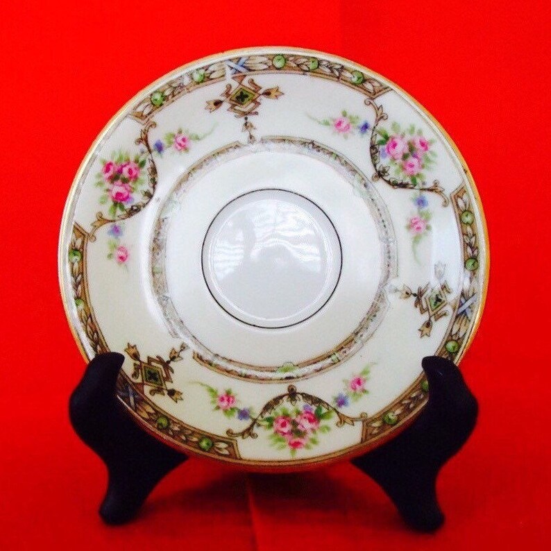 May include: A vintage porcelain saucer with a cream-colored base, adorned with pink rose bouquets and green and gold trim. The saucer has a central recessed area and is displayed on a black stand against a red background.