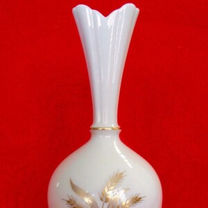 May include: A white, elegant vase with a flared top and a slender neck. The vase features a gold band near the top and a gold wheat design on the lower portion. The background is a solid red.