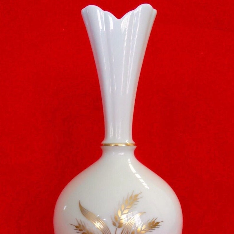 May include: A white, elegant vase with a flared top and a slender neck. The vase features a gold band near the top and a gold wheat design on the lower portion. The background is a solid red.